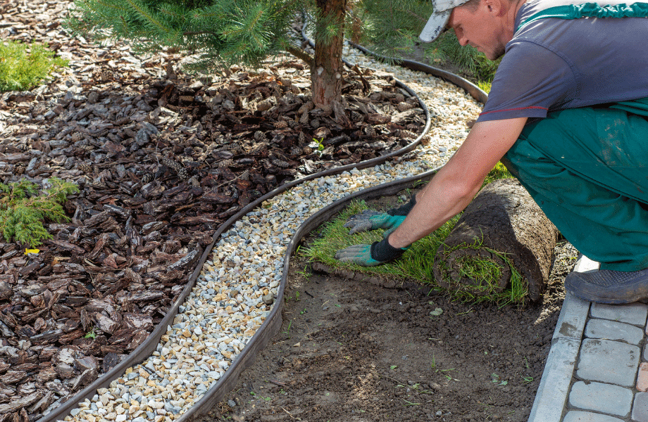 worker in green pants, grey shirt and gloves, laying blue couch turf between a section of pebbles and a section of paving