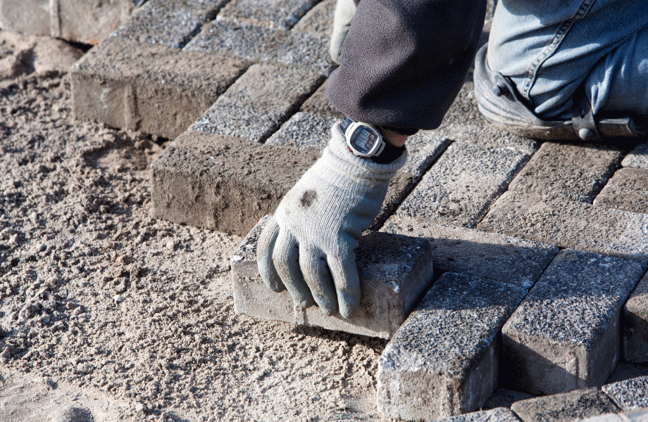 kneeling person with gloved hands laying rectangular grey pavers on prepped dirt for types of pavers