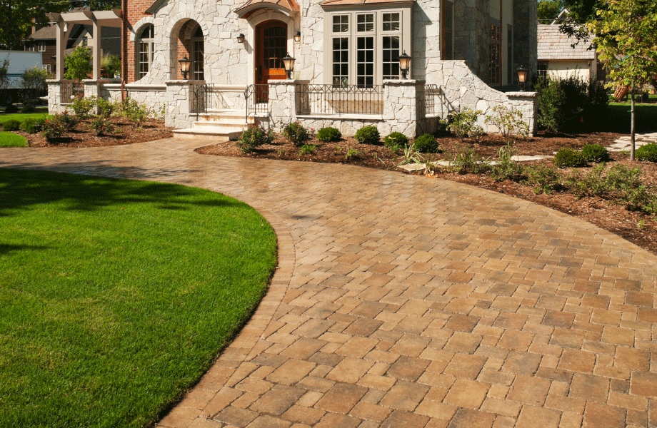 image of a paved driveway bordered by grass and garden beds leading to a white stone home for types of pavers