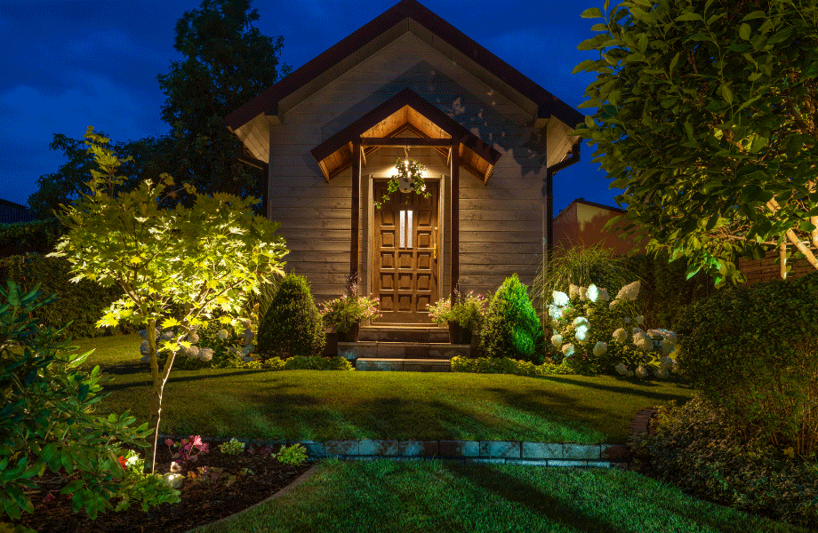 small home with pitched roof and a lit front porch, grass in front with beautiful garden beds with trees, bushes and flowers for rustic garden ideas