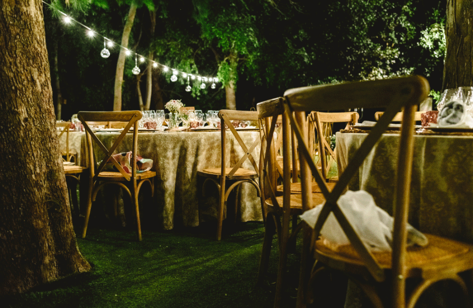 night garden scene with tables with nude tablecloths and wooden chairs, fairy lights and surrounded by trees 