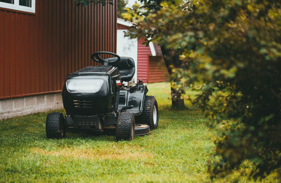 driving lawn mower sitting on a grassy lawn next to a red wooden house with a bush in the foreground