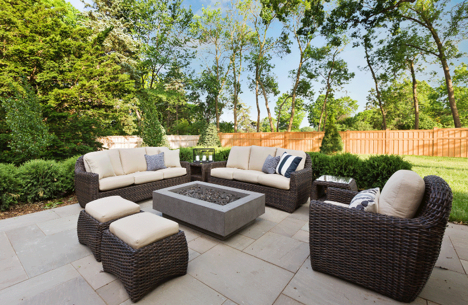 patio area with wicker lounge set and a gray rectangular firepit in the middle next to a green lawn, trees and a brown fence for outdoor entertaining space ideas